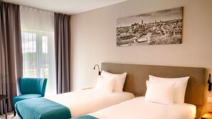 Habitación de hotel con dos camas, una silla, un escritorio y un cuadro de un paisaje urbano en la pared.
