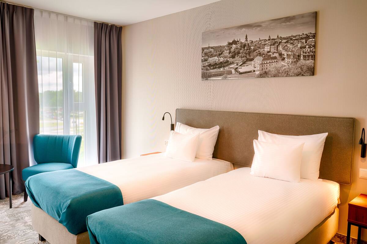 Une chambre d’hôtel avec deux lits, une chaise, un bureau et un tableau représentant un paysage urbain accroché au mur.