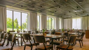 Un comedor amplio con varias mesas y sillas, ventanas grandes con cortinas y plantas en macetas.