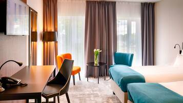 Habitación de hotel con una cama, un escritorio con teléfono, una silla naranja, una silla azul, una mesita con un florero y un ventanal grande con cortinas.