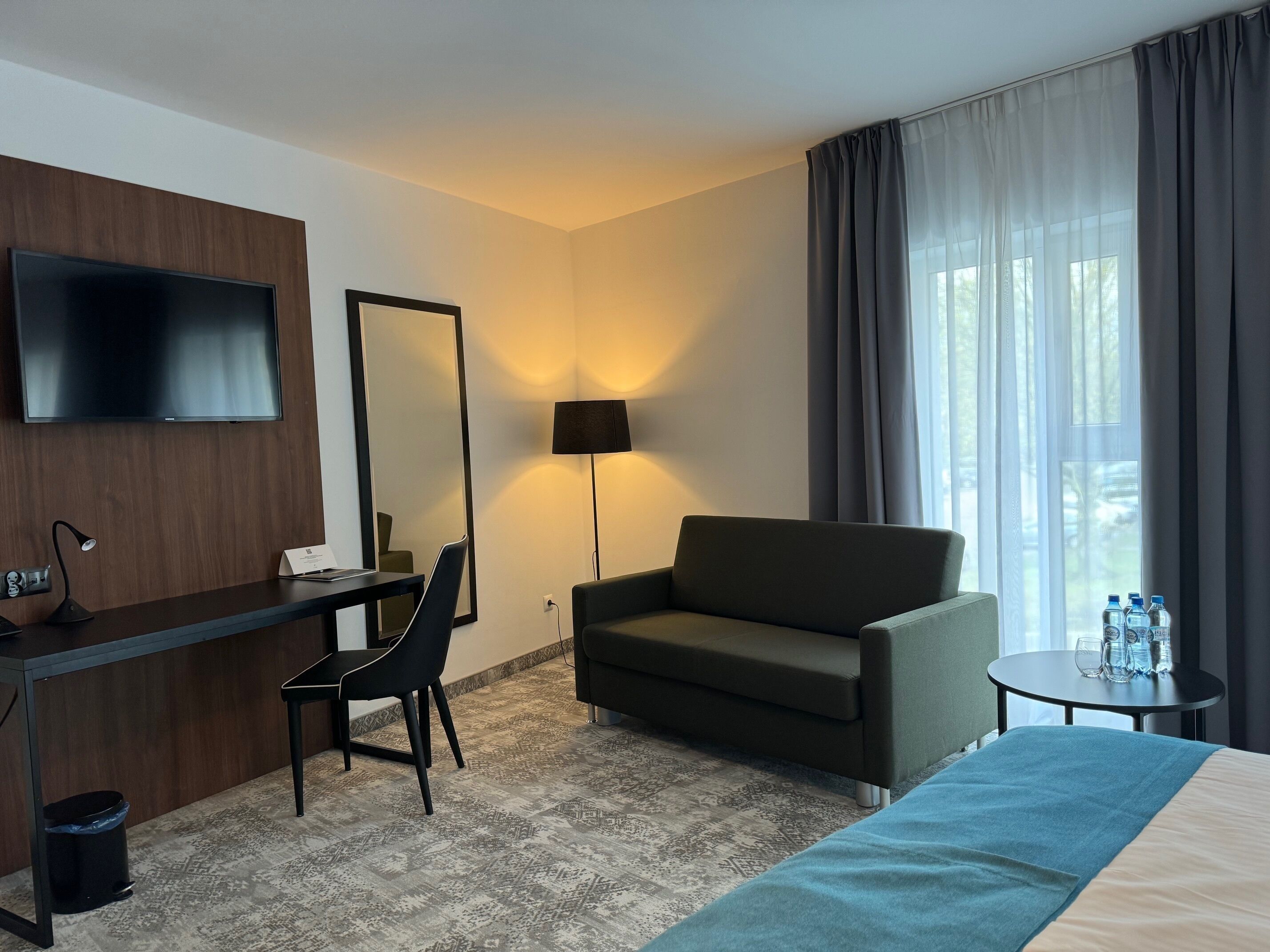 Habitación de hotel con cama, escritorio con silla, sofá, televisor, lámpara y ventana con cortinas.