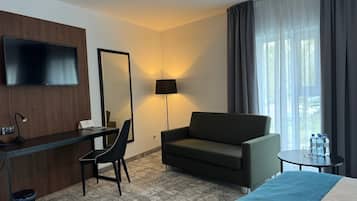 Habitación de hotel con cama, escritorio con silla, sofá, televisor, lámpara y ventana con cortinas.