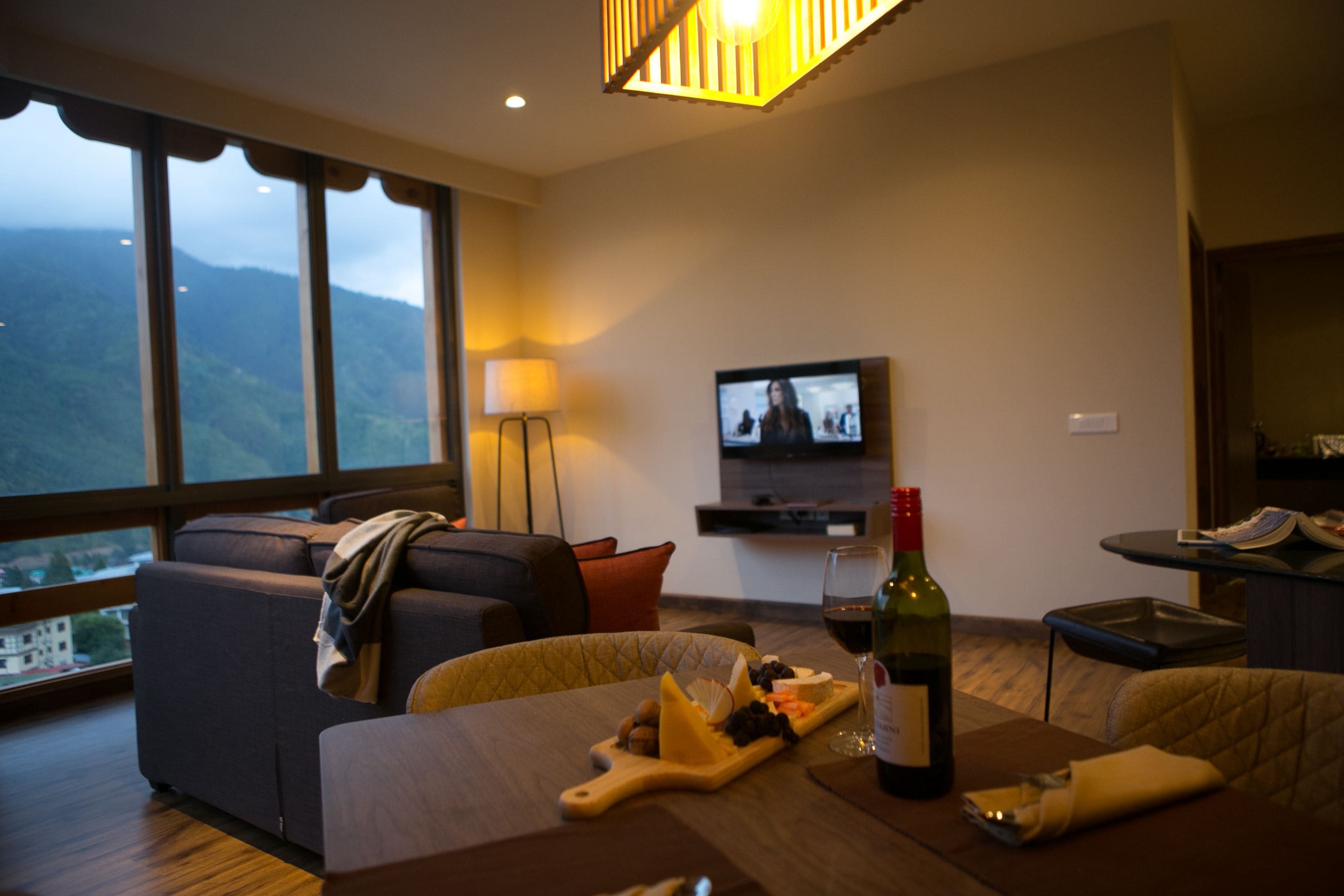 Una sala de estar moderna con un ventanal grande, un televisor de pantalla plana, una botella de vino y una tabla de quesos.