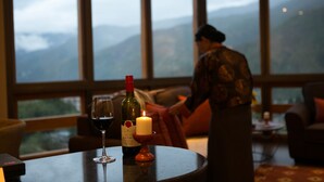 A table with a wine bottle and glass, a candle, and a view of mountains through large windows. - The Pema by Realm (Thimphu)