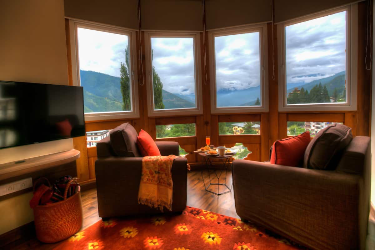 A cozy living room with a large TV, comfortable seating, a small table with a drink, and a view of mountains through large windows.