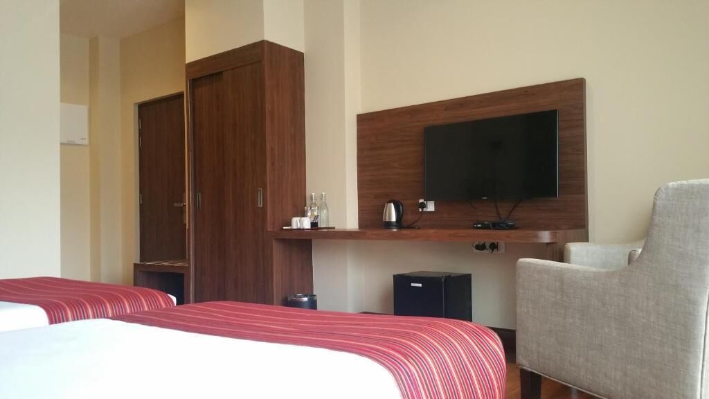 A hotel room with two beds, a TV mounted on the wall, a grey armchair, and a small table with a kettle and cups.