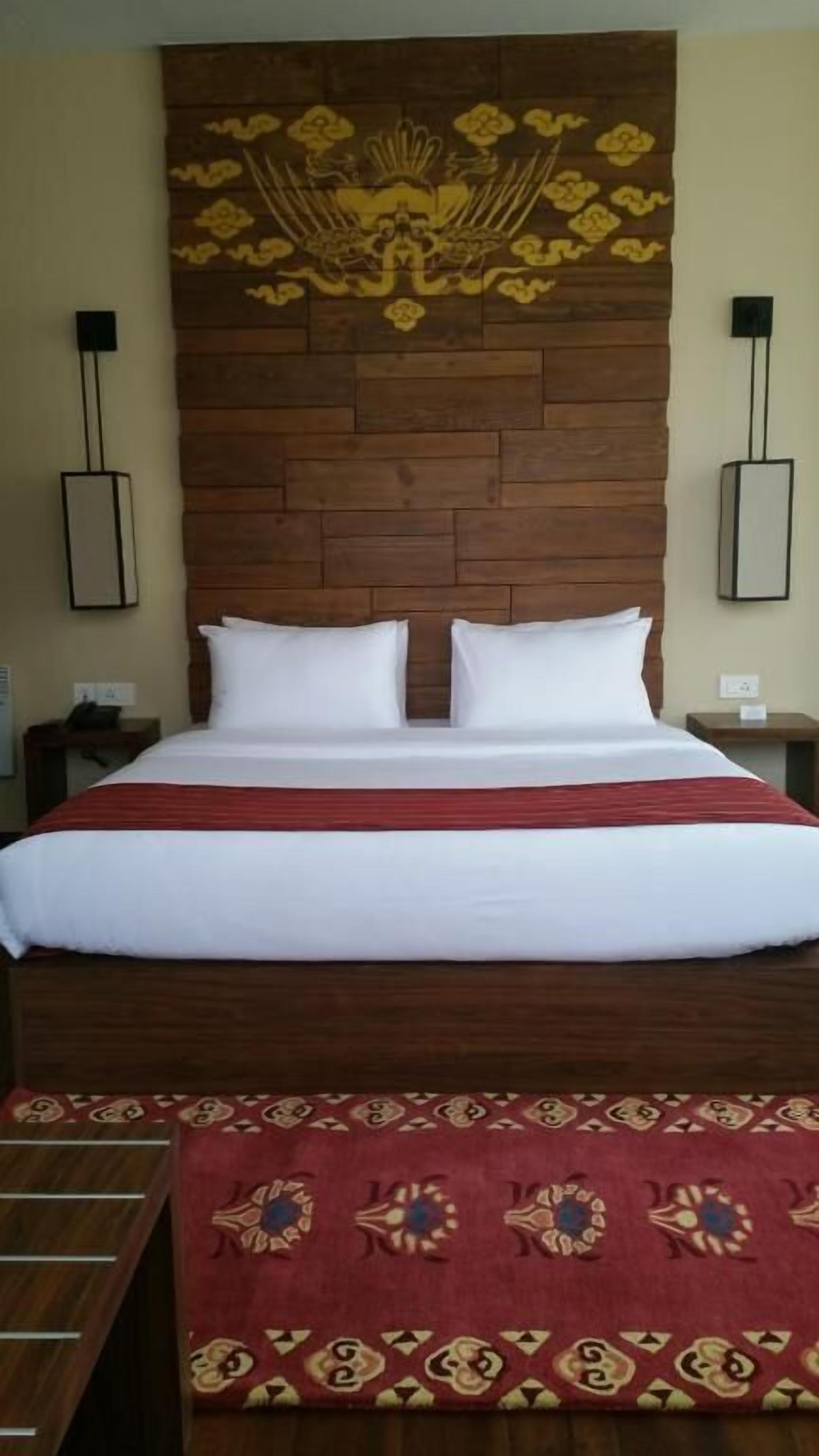 A hotel room with a wooden headboard, two white pillows, a red bedspread, and a patterned red rug.
