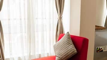 Un sillón rojo con un cojín, una mesita redonda con una taza y un platillo, y un refrigerador en una habitación con cortinas.