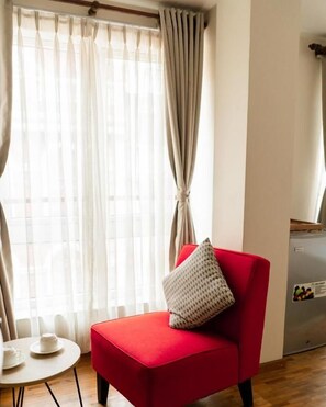 A red armchair with a cushion, a small round table with a cup and saucer, and a refrigerator in a room with curtains. - Laxmi Niwas (Lalitpur)