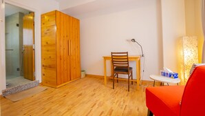 Una habitación con un armario de madera, una mesa con una silla, una mesita con una lámpara y un sofá rojo.