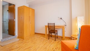 Una habitación con piso de madera, un escritorio con silla, un armario y una mesita de noche con lámpara.