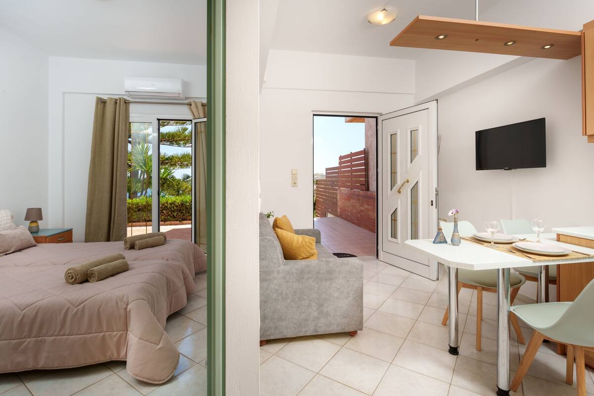 A modern hotel room with a bed, a sofa, a dining area with a table and chairs, and a TV mounted on the wall.