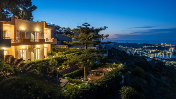A modern multi-story building with balconies, surrounded by lush greenery and overlooking a coastal town at dusk. - FnK Apartments (Malevizi)
