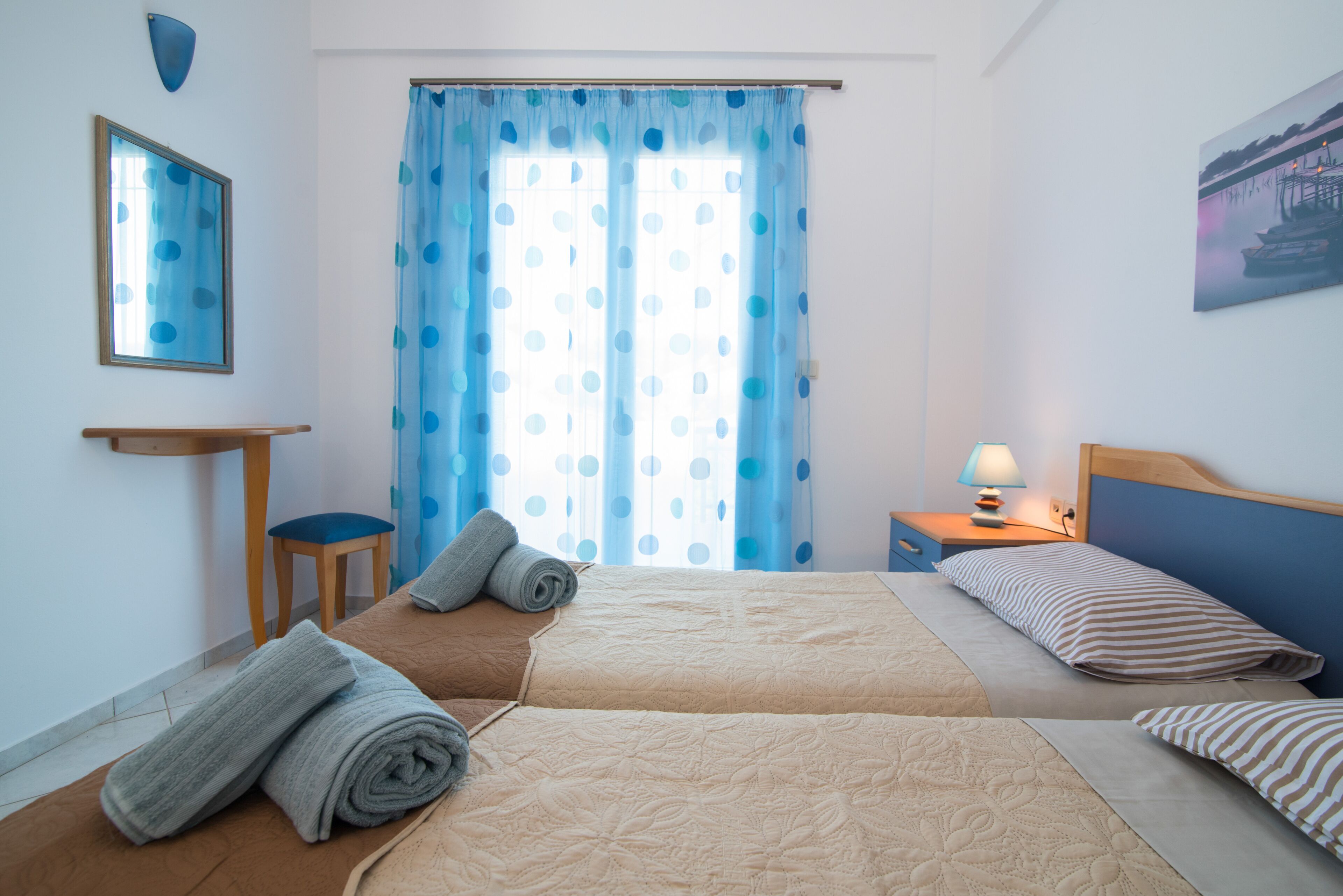A hotel room with two beds, a small table, a chair, a lamp, and a painting on the wall.