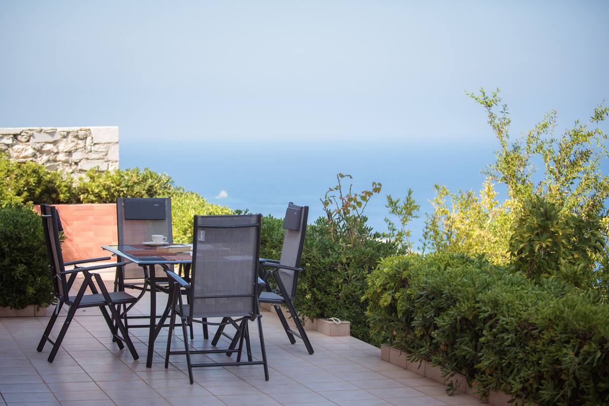 An outdoor patio with a table and chairs, a stone wall, and a view of the sea.