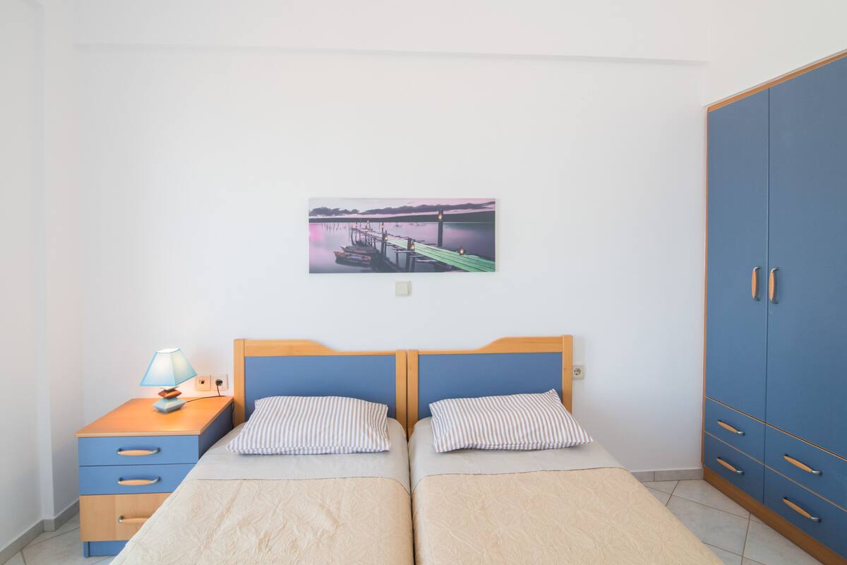 A bedroom with two beds, a nightstand, a wardrobe, and a wall-mounted picture of a dock.