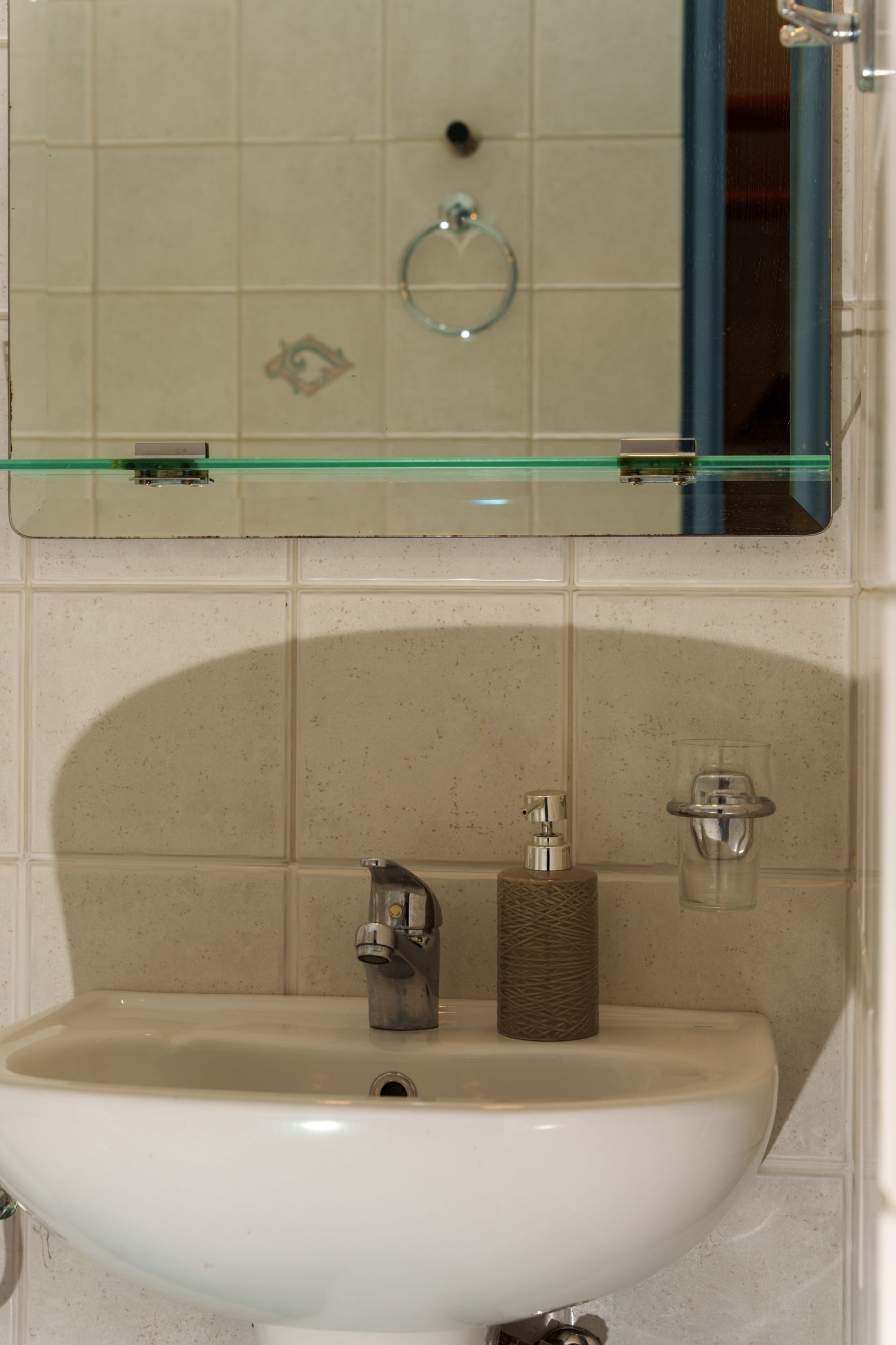 A bathroom with a white sink, a glass shelf above it, and a mirror reflecting a towel ring and a door.