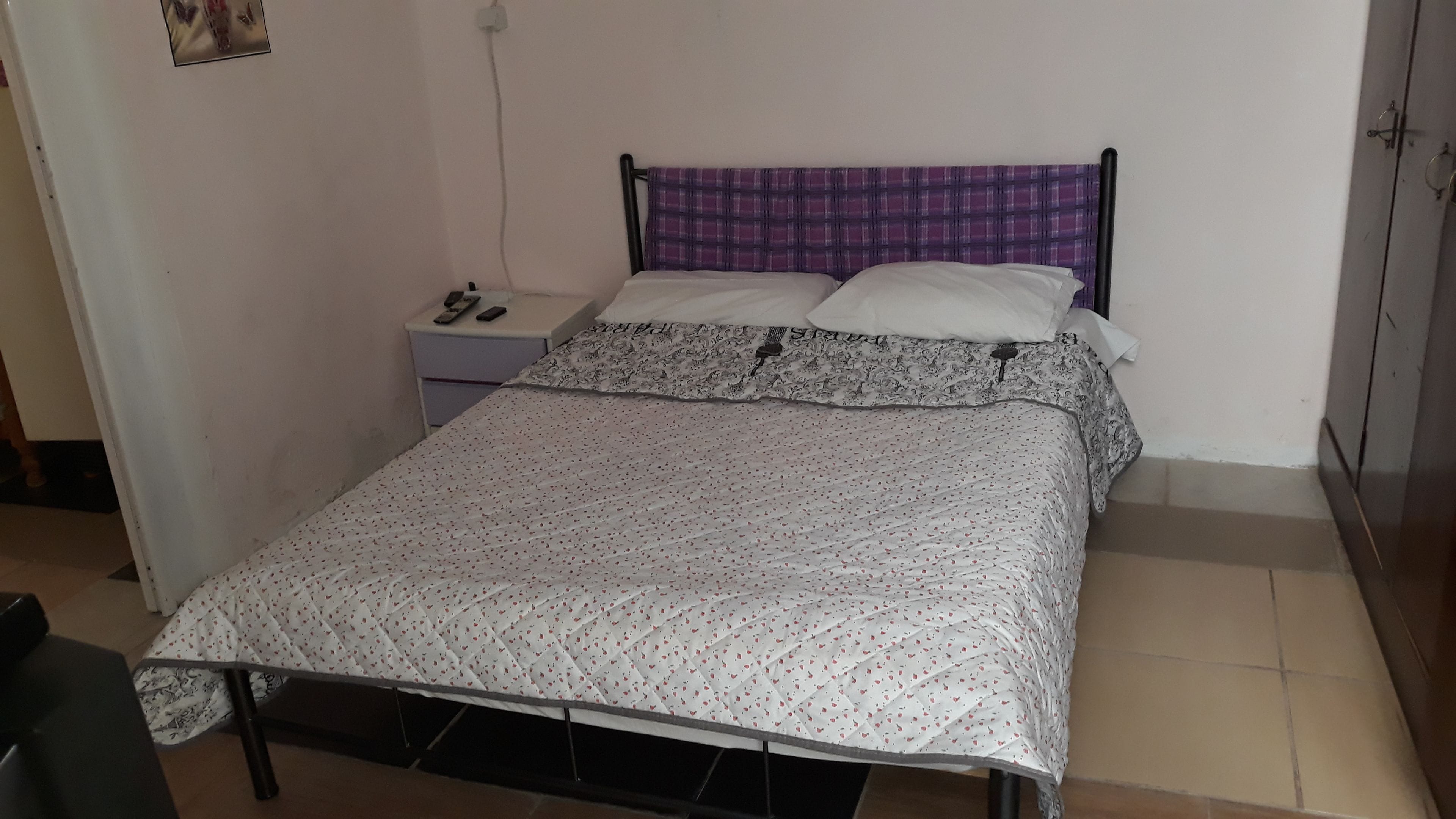 A single bed with a patterned bedspread, a nightstand with a phone and remote controls, and a purple headboard.