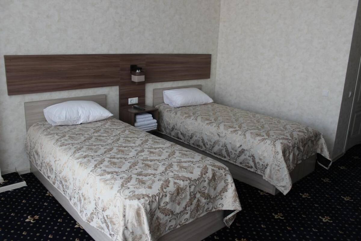 A hotel room with two single beds, each with a wooden headboard and patterned bedspreads. There is a nightstand with a lamp and a folded towel.