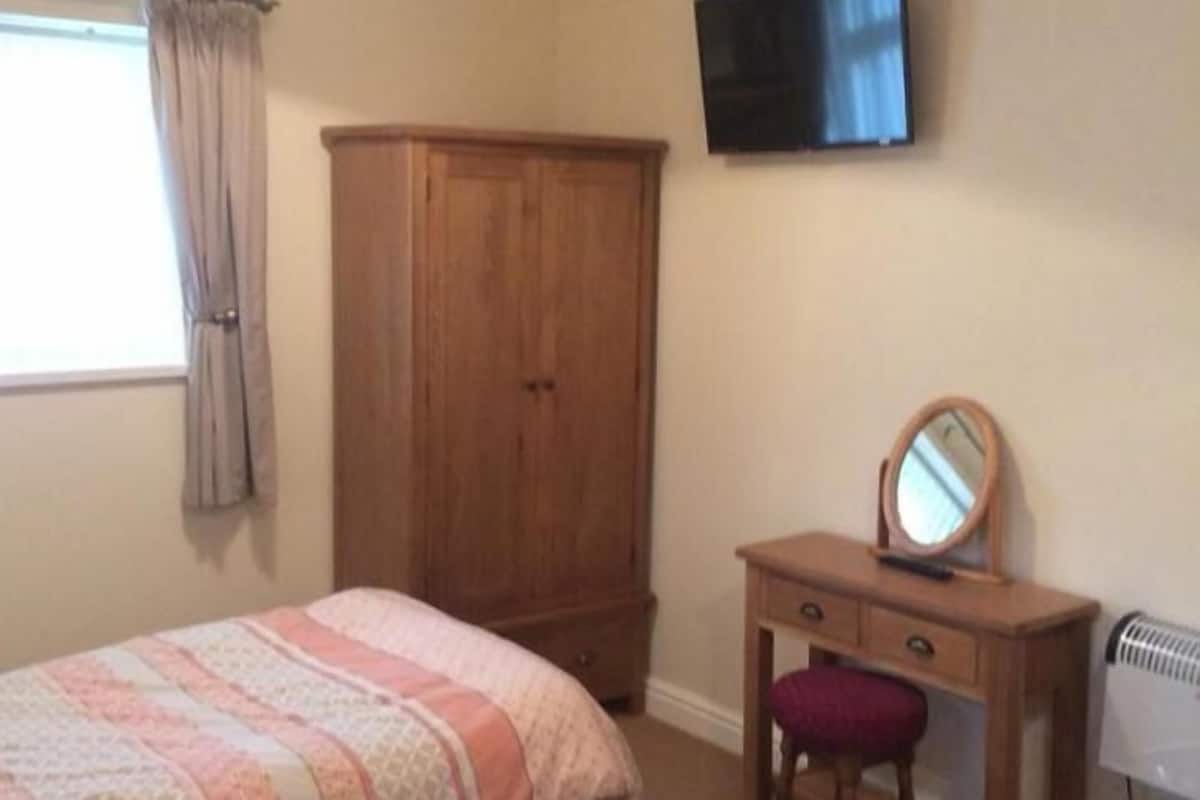 A bedroom with a bed, a wooden wardrobe, a dresser with a mirror, and a wall-mounted TV.
