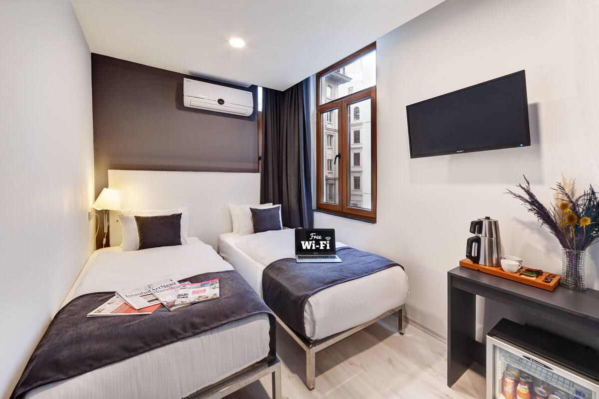 A hotel room with two beds, a TV, an air conditioner, a window with curtains, and a minibar.