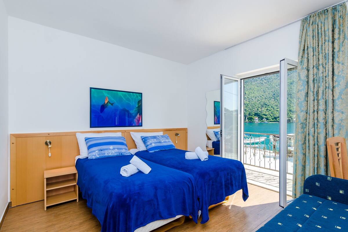 A hotel room with two beds, a wooden headboard, a painting, and a balcony with a view of the sea.