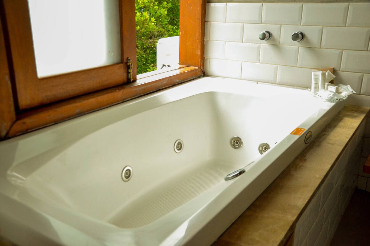 Une grande baignoire rectangulaire dotée de plusieurs robinets, une fenêtre au-dessus et un mur recouvert de carreaux.