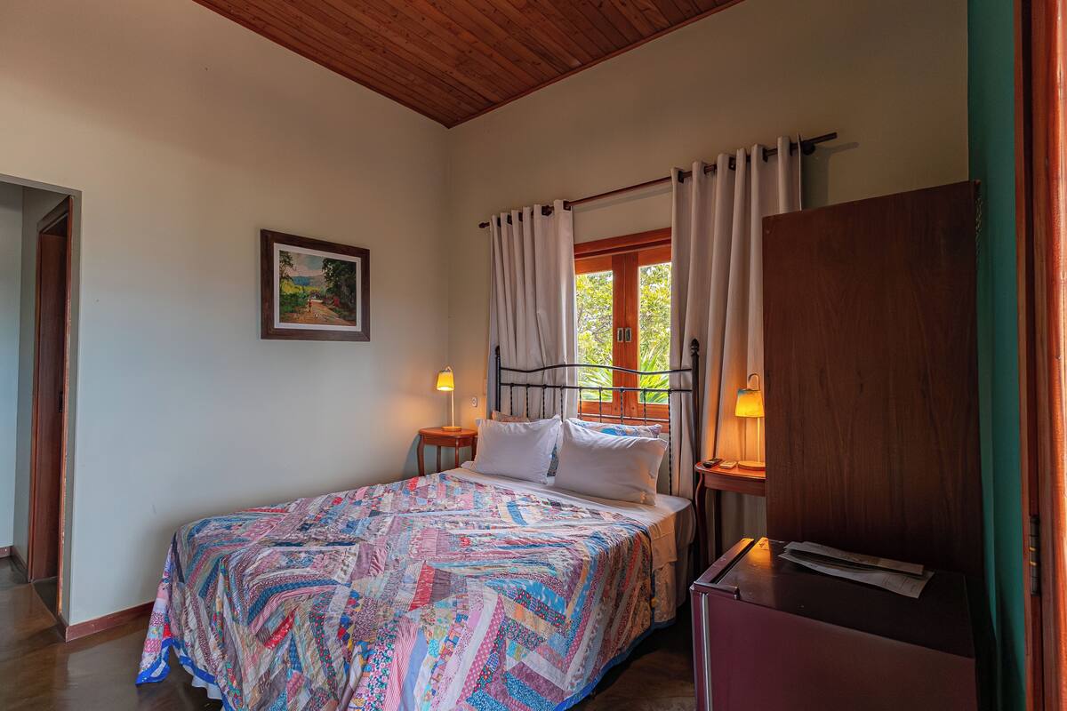 Une chambre à coucher avec un lit, une table de chevet avec une lampe, une armoire en bois, un tableau encadré au mur et une fenêtre avec des rideaux.