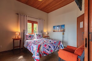 A bedroom with a bed, a window with curtains, a bedside table with a lamp, a painting on the wall, and a door. - Serra do Juá Pousada de Campo (Pirenópolis)