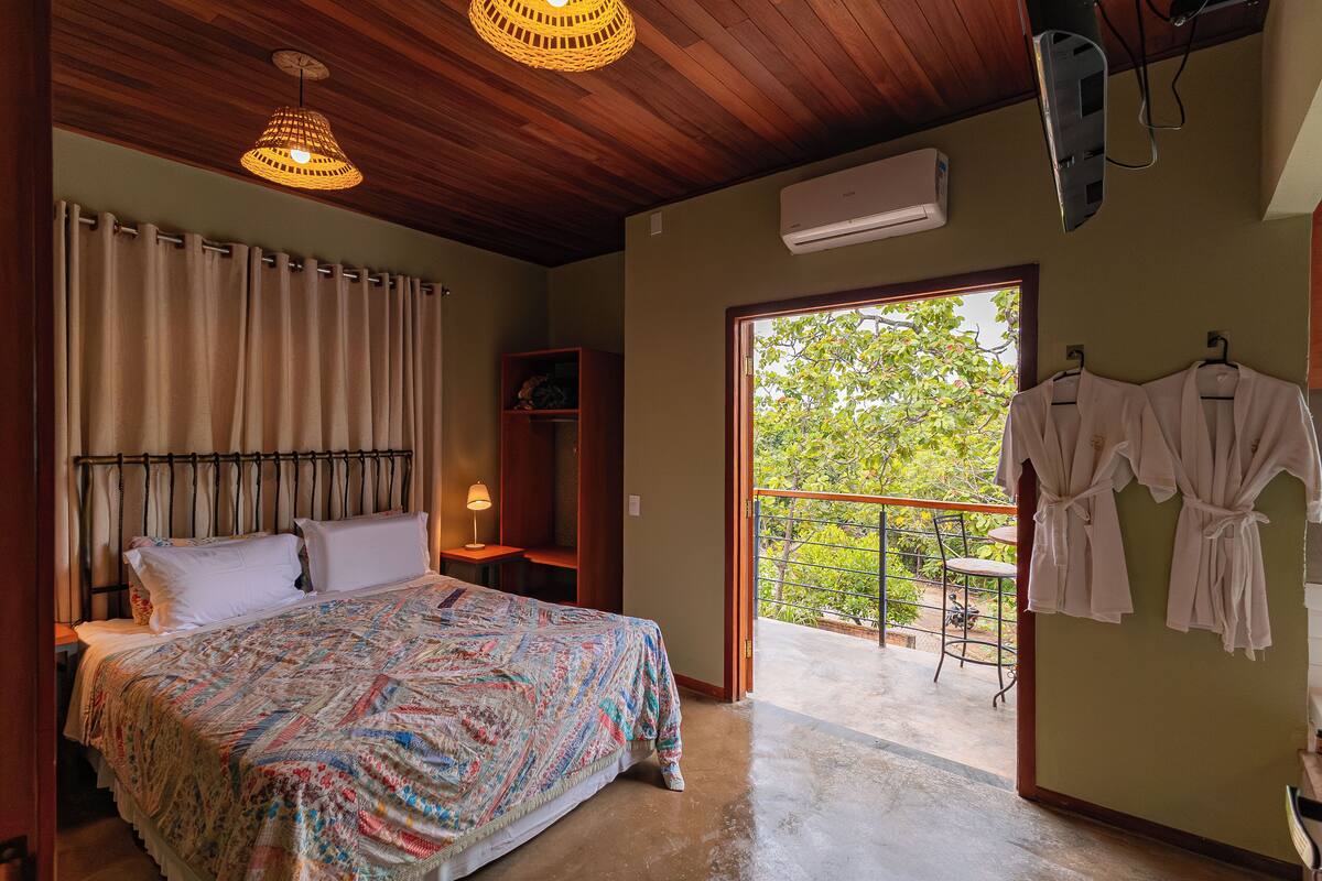 Une chambre à coucher comprenant un lit, des rideaux, une table de chevet, une lampe, une armoire, un balcon avec une table et des chaises, et des vêtements suspendus.