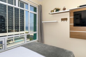 A modern hotel room with a large window, a bed, a TV mounted on the wall, and a small shelf with decorative items.