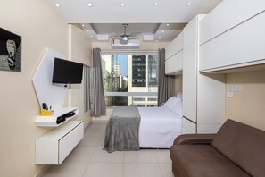 A modern hotel room with a bed, a TV mounted on a white console, a ceiling fan, and a large window with curtains.