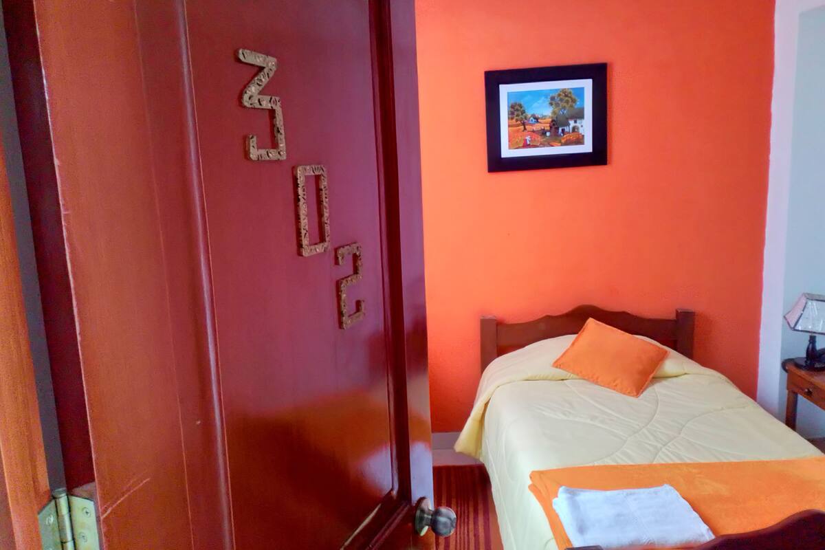 A room with a door marked "302", a bed with an orange pillow, and a framed picture on the wall.