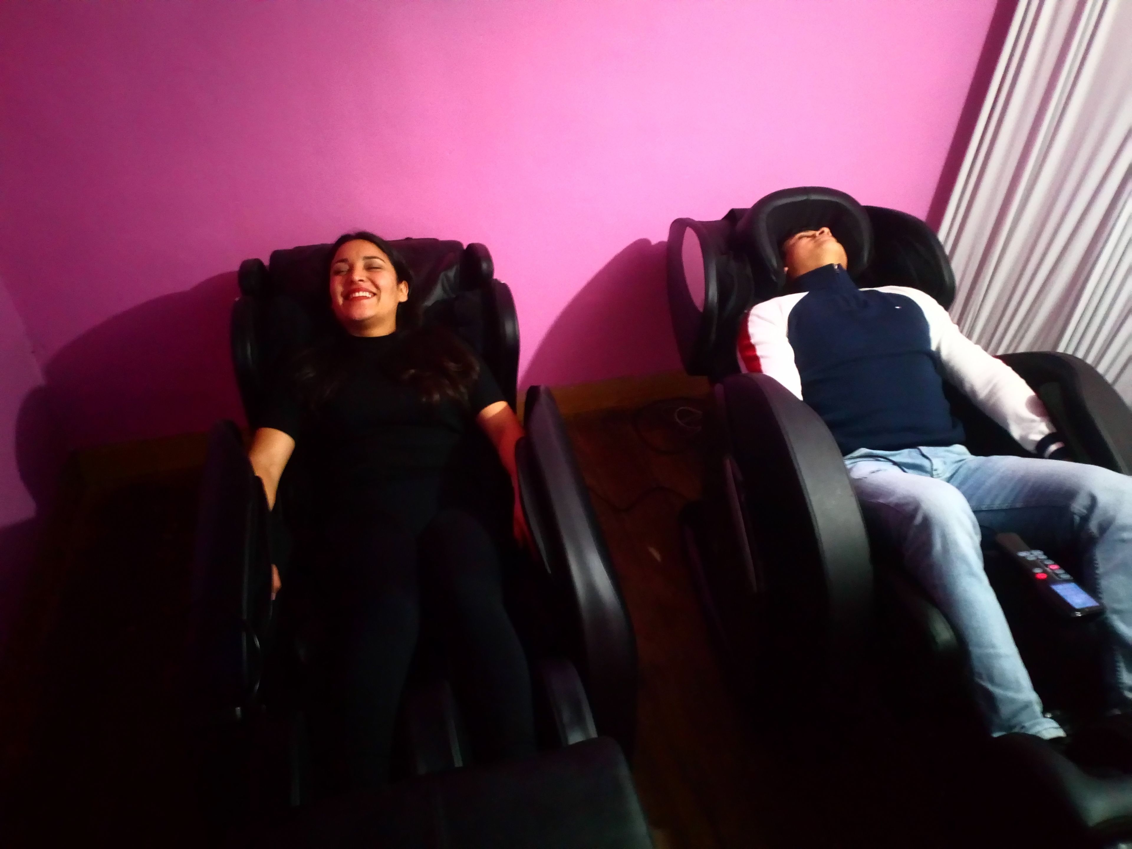 Two people lying in massage chairs in a room with a pink wall.