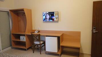 Quarto compacto com escrivaninha de madeira, uma cadeira, um pequeno frigorífico e televisão na parede.