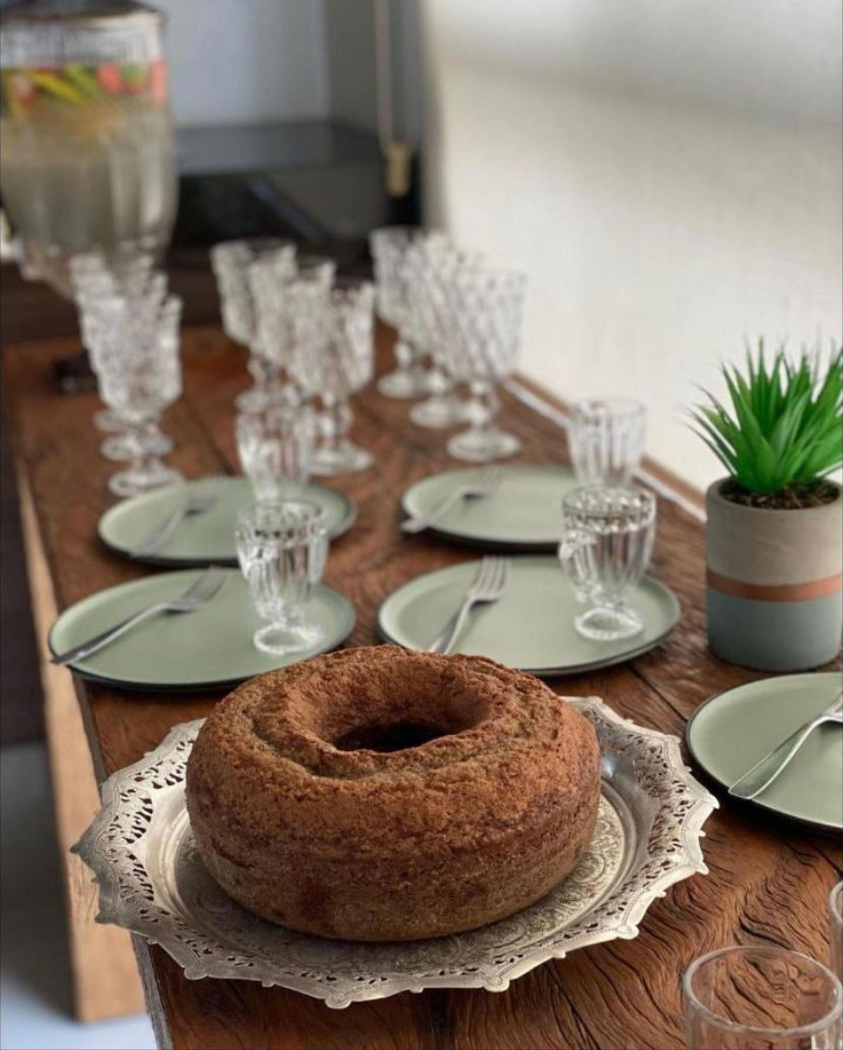 Une table en bois dressée pour un repas, avec un gâteau en guise de centre de table, de la verrerie transparente et une plante en pot.