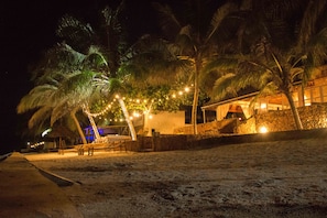 Une plage bordée de palmiers et illuminée de guirlandes lumineuses. On y aperçoit un bâtiment avec un mur en pierre et une structure surmontée d’un toit de chaume.