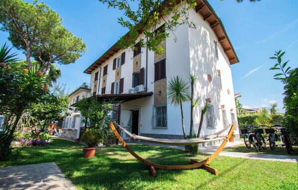 A two-story building with a white facade, multiple windows, and a balcony. There is a hammock in the front yard, and several bicycles are parked nearby. - Hotel Villa Giada (Massa)