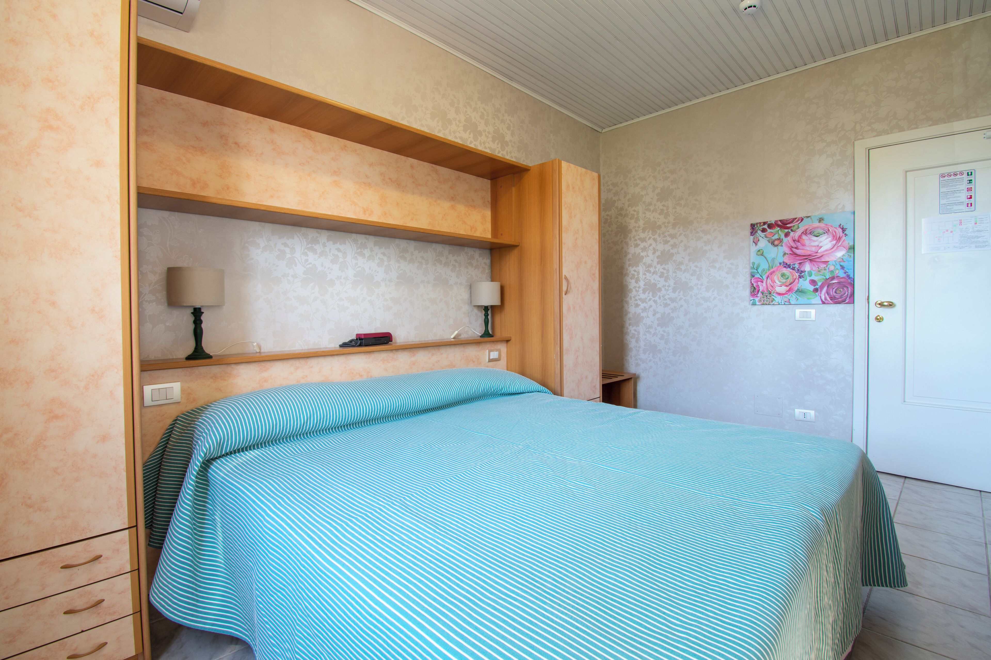 Une chambre à coucher avec un lit, une étagère sur laquelle se trouve une lampe, une porte et un mur orné d’un tableau représentant des motifs floraux.