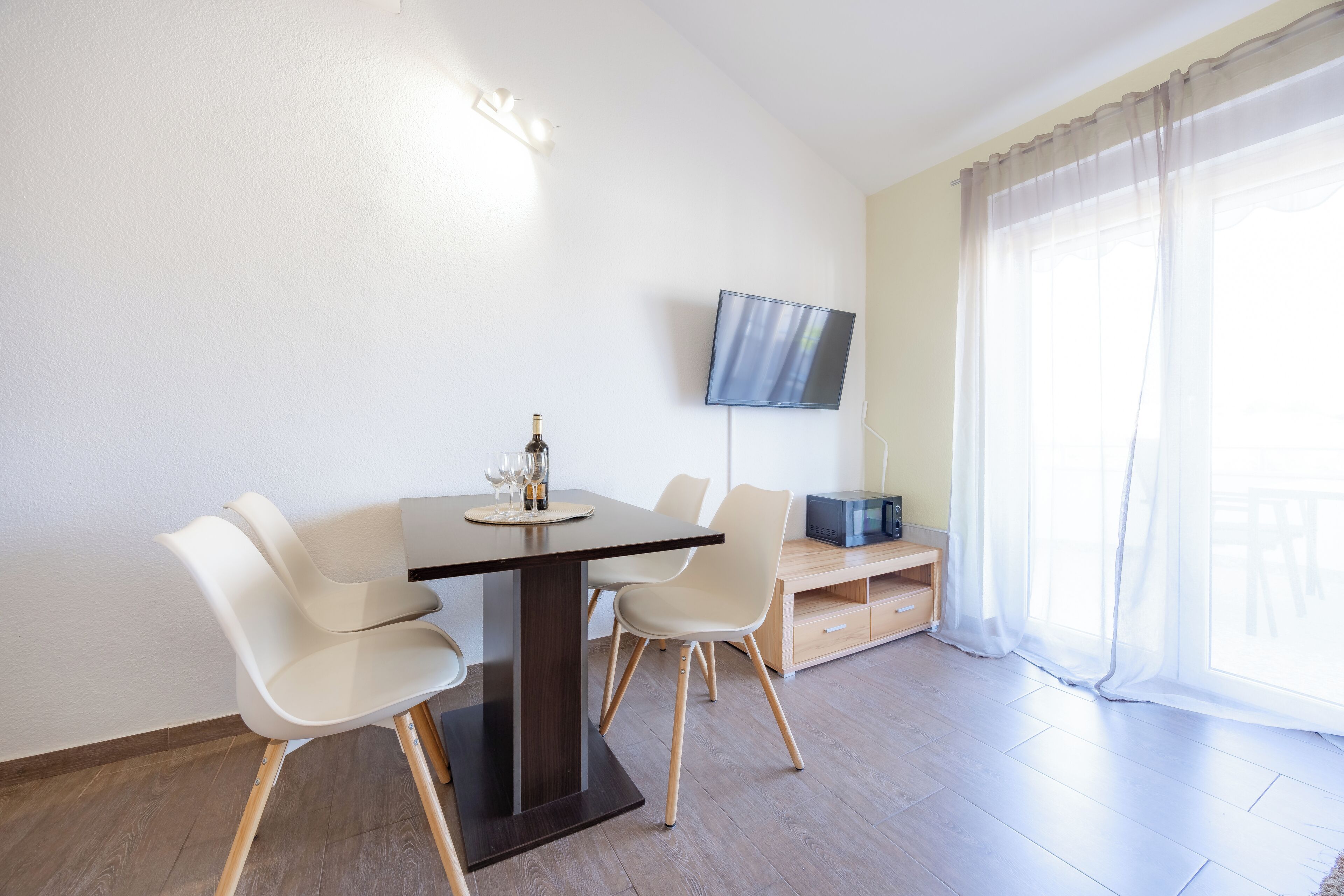 Una habitación moderna con una mesa de comedor, sillas, un televisor de pantalla plana y un ventanal con cortinas translúcidas.
