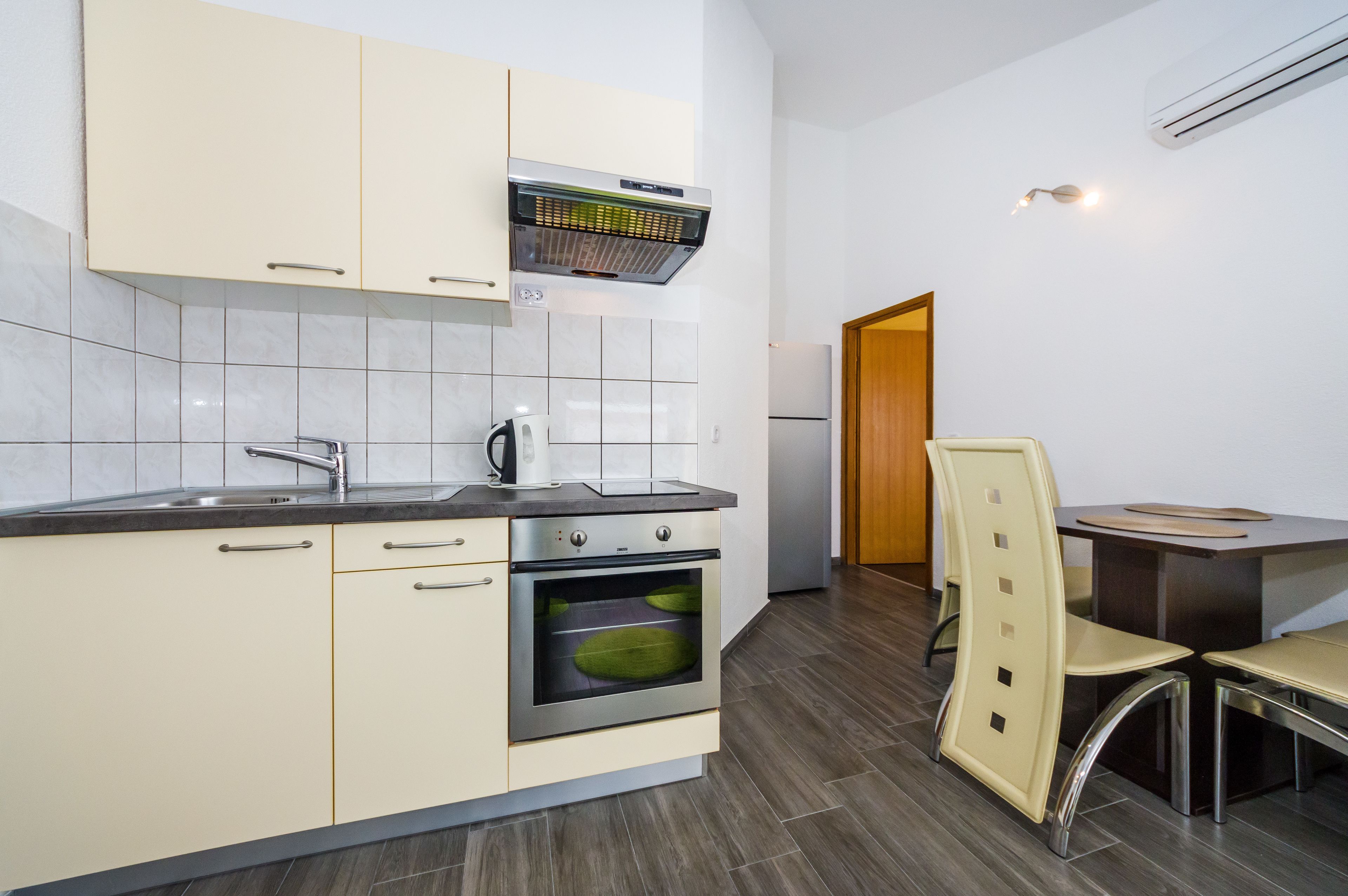 Une cuisine moderne avec des armoires blanches, un four encastré, une hotte de cuisinière en acier inoxydable et un coin repas avec une table et des chaises.