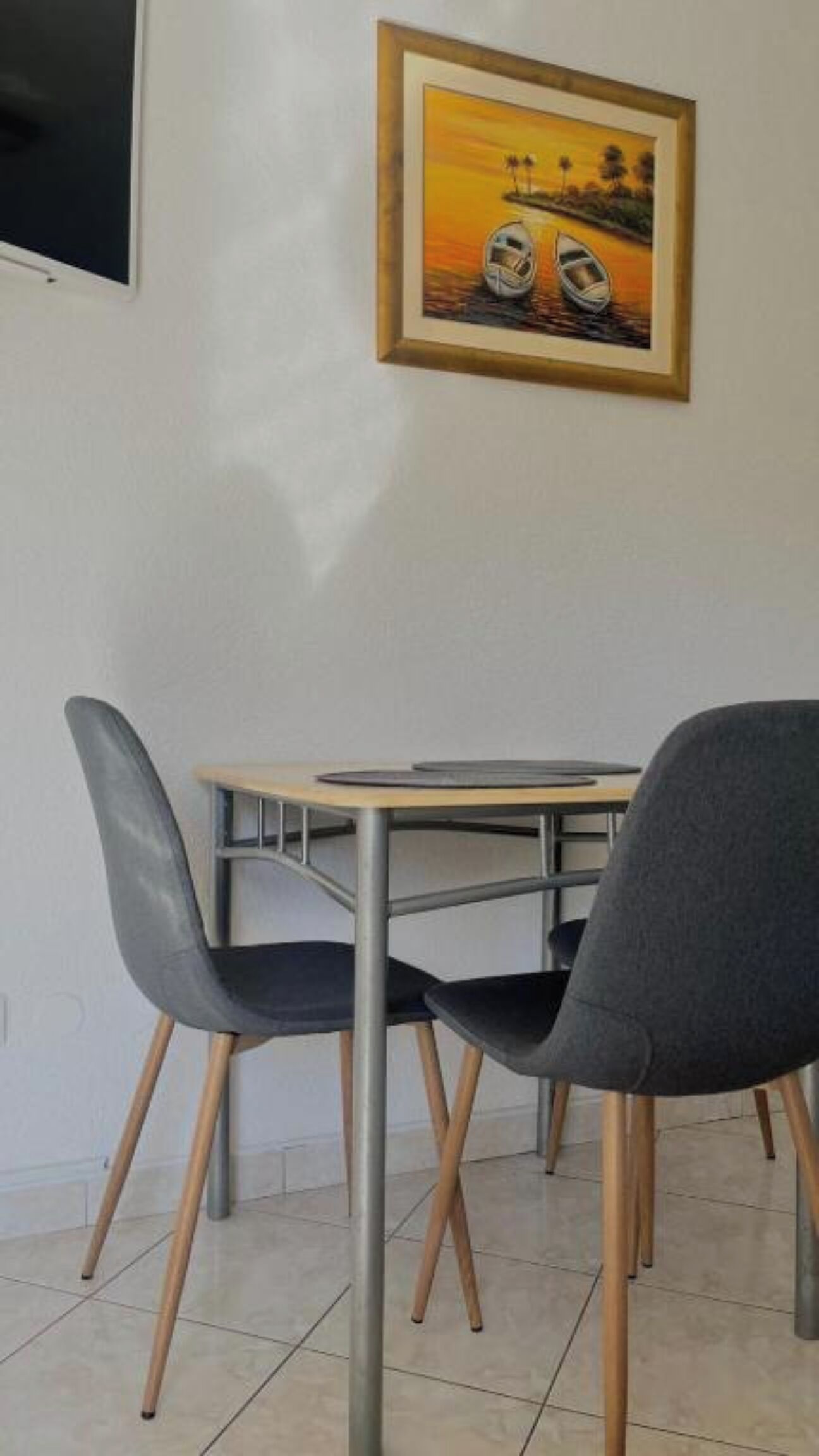 Un comedor con una mesa y cuatro sillas, un cuadro enmarcado en la pared y suelo de baldosas.