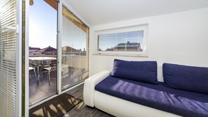 Una sala de estar moderna con un sofá blanco y cojines azules, un ventanal corredizo que da a un área exterior con una mesa y sillas, y una ventana con persianas.