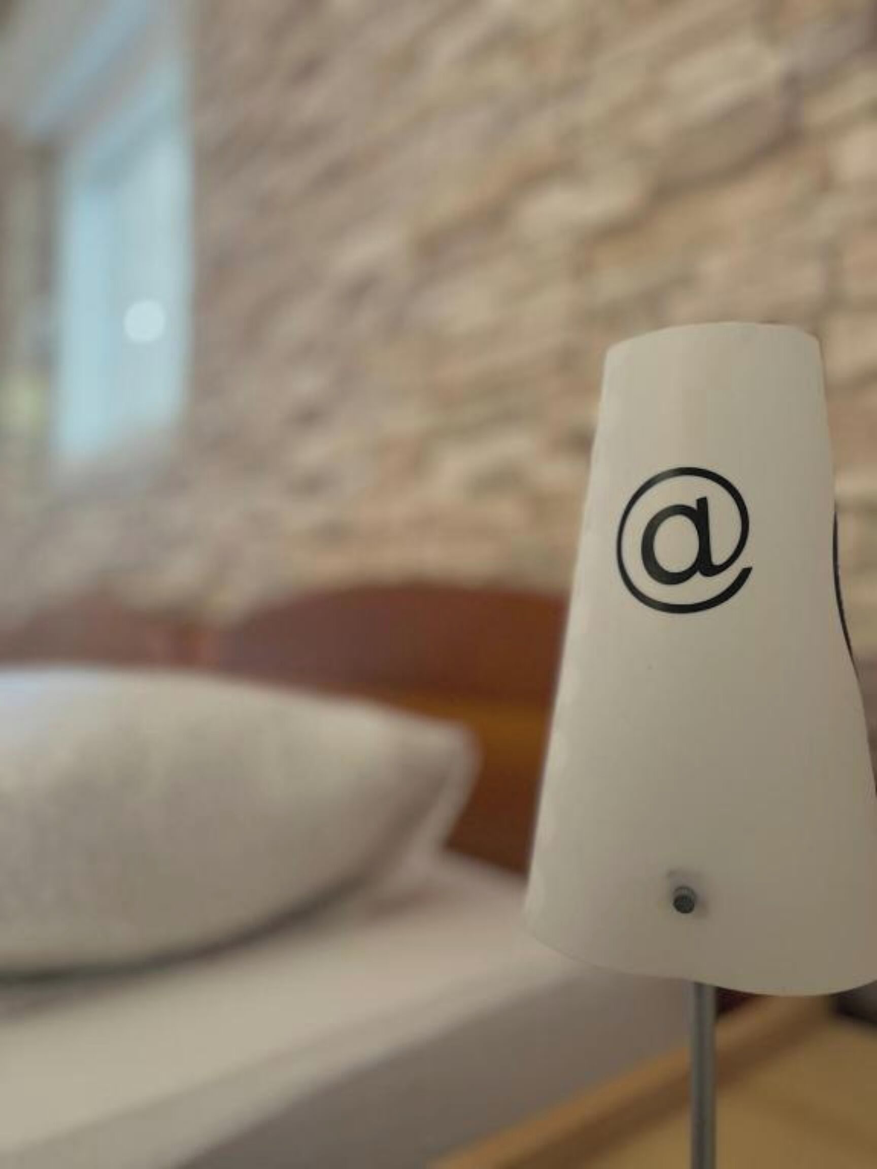 A white lamp with an '@' logo on a bedside table in a room with a brick wall and a window.