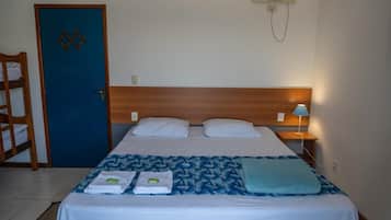 Quarto de hotel com cama, porta azul, cabeceira de madeira, mesa de cabeceira com abajur e ar-condicionado fixado na parede.