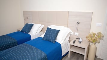 Habitación de hotel con dos camas, cada una con ropa de cama azul y almohadas blancas, una mesita de noche con teléfono y control remoto, y un jarrón con flores.