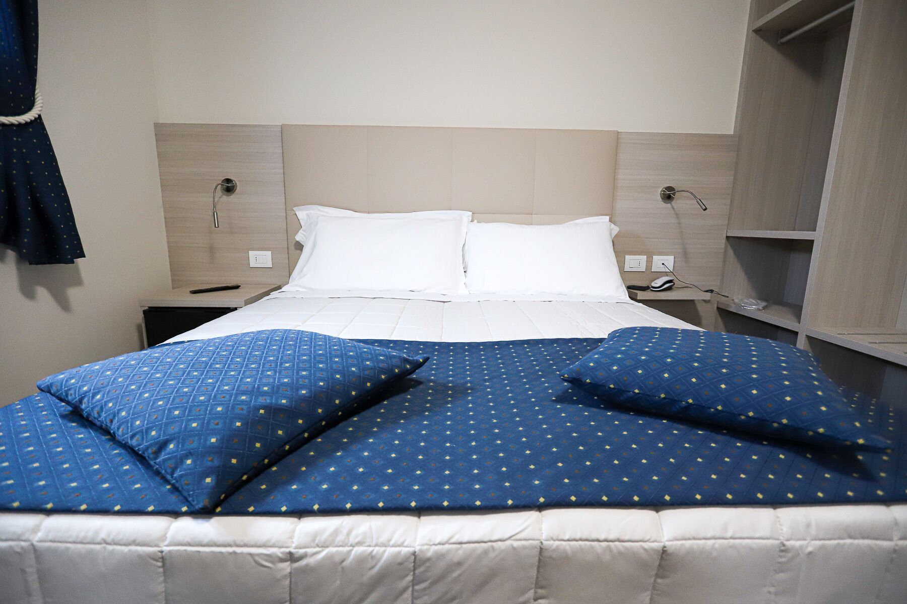 Quarto de hotel com uma cama grande, duas almofadas brancas e um cobre-leito azul com padrão. Há duas mesas de cabeceira com luminárias e uma tomada em cada uma.