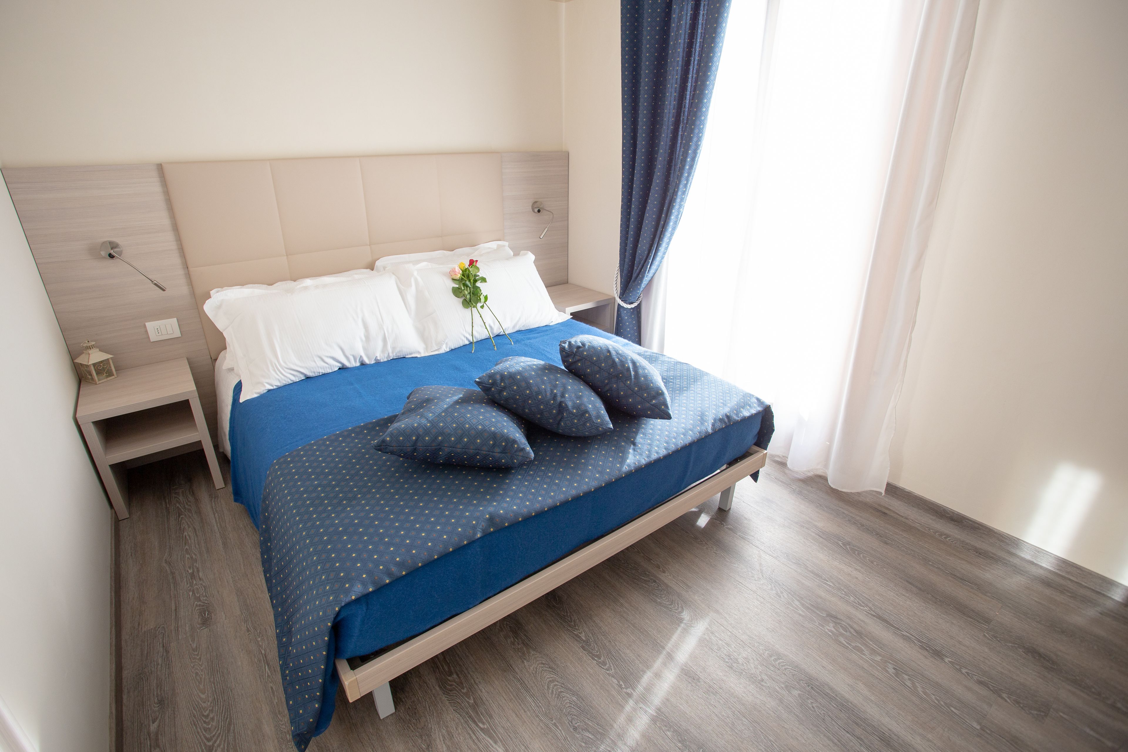Una cama bien hecha con una colcha azul y almohadas, una mesita de noche con una lámpara, y una ventana con cortinas.
