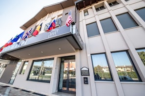Entrada de hotel con fachada moderna, varias banderas y puerta de vidrio.