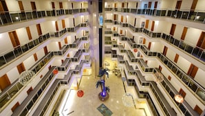 Un vestíbulo de hotel de varios niveles con una escalera central, barandillas de vidrio e iluminación decorativa.
